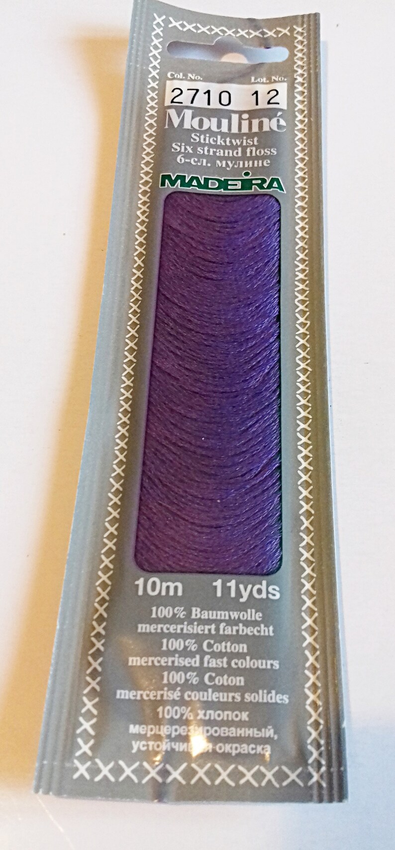 Puede incluir: Un ovillo de hilo de bordar Moulin&eacute; Madeira de color morado, etiquetado con "2710 12" y "Sticktwist Six strand floss 6-сл. мулине". El embalaje tambi&eacute;n indica "10m 11yds" y enumera el contenido de la fibra como 100% algod&oacute;n mercerizado.