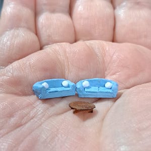 May include: Two miniature light blue sofas with small white cushions and a tiny round brown table. These dollhouse-scale furniture pieces are crafted with a textured finish and are shown resting on a palm to demonstrate their small size.