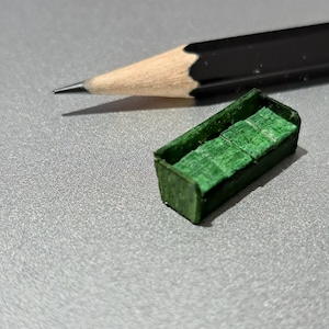 May include: A black pencil with a sharpened tip and a small, green rectangular object. The green object appears to be a small block of wood or a similar material, possibly a component or a miniature item.