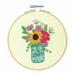 Puede incluir: Arte de bordado en aro que presenta un tarro de cristal azul lleno de un ramo de flores de colores, incluyendo un girasol, flores rojas y rosas, y margaritas blancas. El tarro tiene la palabra "Love" escrita en él.