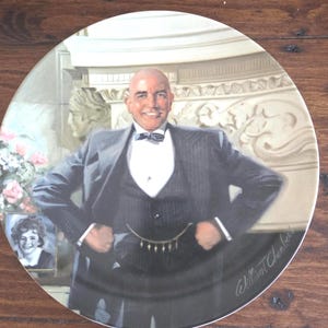 May include: A decorative plate with a portrait of a man in a suit and bow tie. The plate has a gold rim and a signature in the bottom right corner. The man is smiling with his hands on his hips.