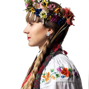 May include: A woman wearing a traditional white blouse with colorful floral embroidery. She is wearing a wreath of flowers on her head and a red and black sash around her neck. Her hair is braided and she is wearing pearl earrings.