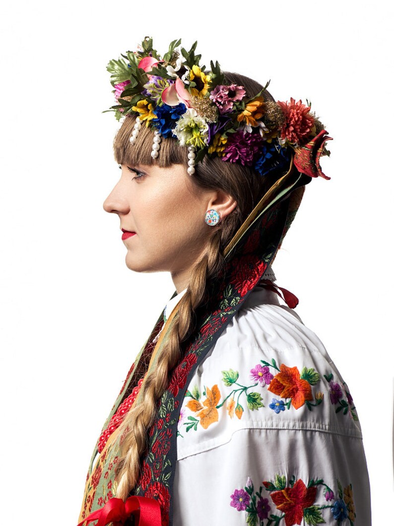 May include: A woman wearing a traditional white blouse with colorful floral embroidery. She is wearing a wreath of flowers on her head and a red and black sash around her neck. Her hair is braided and she is wearing pearl earrings.
