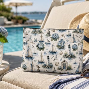 Puede incluir: Un estuche rectangular con cremallera que presenta un estampado náutico repetitivo sobre un fondo blanco. El diseño incluye veleros azules, faros, gaviotas, conchas marinas y racimos de hortensias azules por toda la superficie.