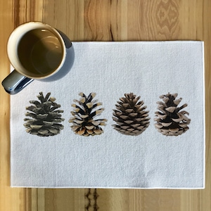 May include: A white linen placemat with four brown pine cones arranged in a row. The pine cones are different sizes and shapes.