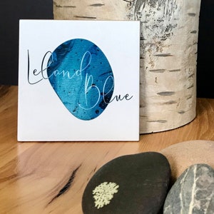 May include: White square sign with the text "Leland Blue" in black cursive script. A blue abstract shape is behind the text.