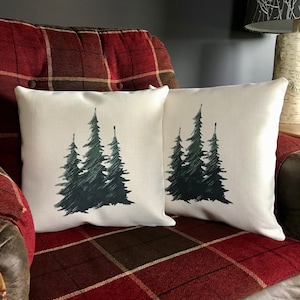May include: Two white throw pillows with a forest scene design. The pillows feature a dark green silhouette of three pine trees on a white background. The pillows are sitting on a red and brown plaid couch.