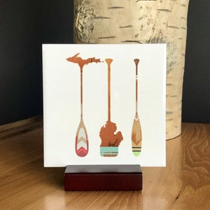 May include: Three wooden paddles with different colored designs. The paddles are painted in shades of brown, red, green, and white. The paddles are arranged vertically on a white background.
