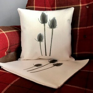 May include: Two white throw pillows with a design of brown thistle flowers on the front. The pillows are on a red and brown plaid couch.