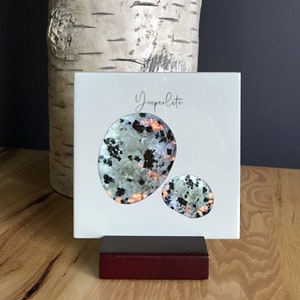 May include: A white square tile with a stand, featuring two Yooperlite stones. The stones are a light green with black and orange flecks. The tile has the word "Yooperlite" written in black cursive.