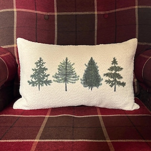 May include: A rectangular, off-white pillow with a textured surface. The pillow features four stylized green Christmas trees in a row. The pillow is resting on a red and brown plaid patterned chair.