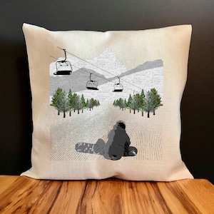 May include: A white throw pillow with a black and white illustration of a snowboarder on a snowy mountain slope. The illustration includes a ski lift with four chairs and a mountain range in the background.