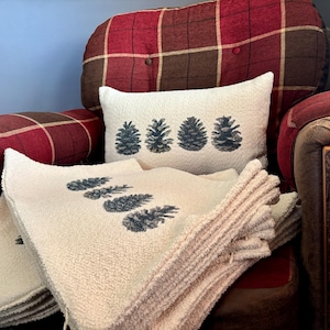 May include: Cream-colored throw pillows and blankets with a repeating pine cone design in dark blue. The pillows and blankets are stacked on a red plaid armchair with a matching plaid headrest.