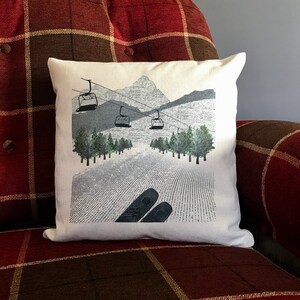 May include: White throw pillow with a graphic of a snowy mountain scene with ski lifts and skiers. The pillow has a red and brown plaid background.