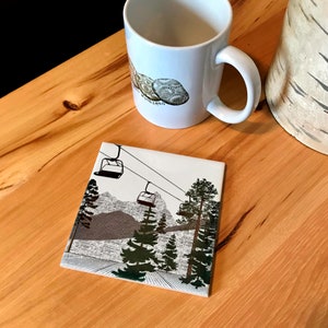 May include: A white ceramic coaster with a black and white illustration of a ski lift with three gondolas and pine trees. The background is a gray and white illustration of a mountain range.