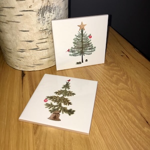 May include: Two white ceramic tiles with a Christmas tree design. The top tile features a green tree with a gold star, a red ornament, and a red cardinal. The bottom tile features a green tree with a red ornament and a red cardinal.