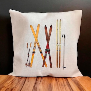 May include: A white pillow with a vintage ski design. The pillow features a pair of skis, ski poles, and a pair of ski boots. The design is in shades of brown, yellow, and red.