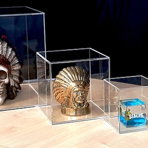 May include: Four clear acrylic cube display boxes of varying sizes. The largest box contains a white skull wearing a red, white and green headdress. The second largest box contains a gold-coloured skull wearing a gold headdress. The third box contains a blue glass cube with the word "SEALIFE" printed on it. The smallest box contains a small red and blue Spiderman figurine with a white spiderweb behind it.