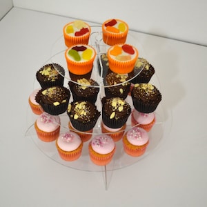 May include: A three-tiered acrylic cupcake stand with pink, chocolate, and fruit-topped cupcakes. The stand is clear and has a circular base.