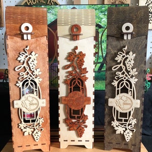 May include: Three wooden wine bottle boxes with intricate laser cut designs. The boxes are stained in different shades of brown and feature a grapevine motif with the word "Wine" on a label.