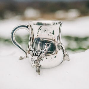 May include: A polished silver-toned metal mug featuring a curved handle and three-dimensional deer head sculptures with prominent antlers attached to the exterior surface of the vessel, creating a decorative and textured metallic finish.