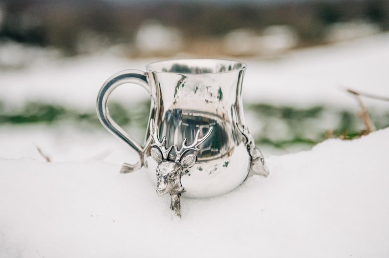 May include: A polished silver-toned metal mug featuring a curved handle and three-dimensional deer head sculptures with prominent antlers attached to the exterior surface of the vessel, creating a decorative and textured metallic finish.