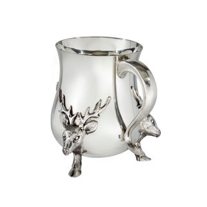 May include: A polished silver-toned mug featuring three dimensional deer head accents that serve as the base and feet of the vessel. The mug has a smooth, reflective surface, a wide rim, and a curved handle extending from the side of the body.