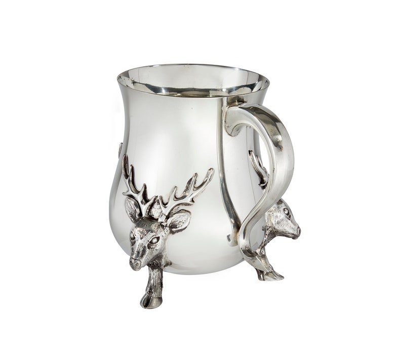 May include: A polished silver-toned mug featuring three dimensional deer head accents that serve as the base and feet of the vessel. The mug has a smooth, reflective surface, a wide rim, and a curved handle extending from the side of the body.