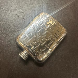 May include: A silver hip flask with a geometric pattern embossed on the surface. The flask has a hinged lid and a small spout.