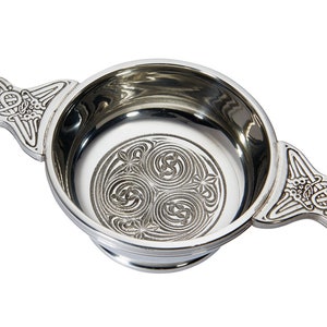 May include: A polished silver-toned metal quaich bowl featuring two side handles with intricate embossed knotwork patterns. The interior base of the bowl displays a circular engraved design consisting of three interconnected spiral motifs.