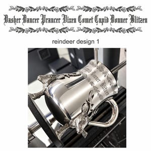 May include: A polished silver-toned metal mug featuring two three-dimensional reindeer heads attached to the sides. The mug is engraved with the text "Dasher Dancer Prancer Vixen Comet Cupid Donner Blitzen" in a decorative font, framed by holly leaves.