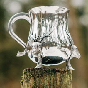 May include: A polished silver-toned metal mug featuring a prominent handle and three-dimensional deer head sculptures integrated into the base. The reflective surface of the vessel displays a curved silhouette and a smooth, metallic finish throughout.