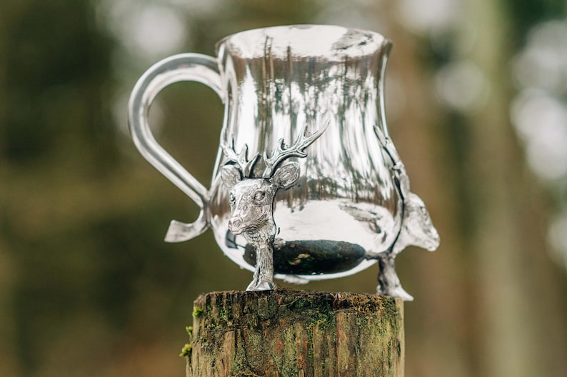 May include: A polished silver-toned metal mug featuring a prominent handle and three-dimensional deer head sculptures integrated into the base. The reflective surface of the vessel displays a curved silhouette and a smooth, metallic finish throughout.