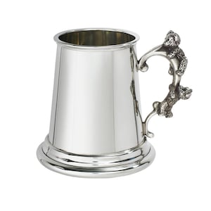 Peut inclure: Une chope en métal argenté poli avec un corps cylindrique lisse et effilé ainsi qu'une base large et arrondie. La poignée présente un design sculptural texturé représentant deux petits animaux empilés aux surfaces détaillées.