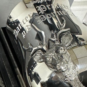May include: A polished silver-toned metal vessel featuring a three-dimensional deer head relief on the front. The surface is engraved with the text Dancer, Prancer, and Donner in a gothic-style font, accompanied by a small pine cone branch motif.