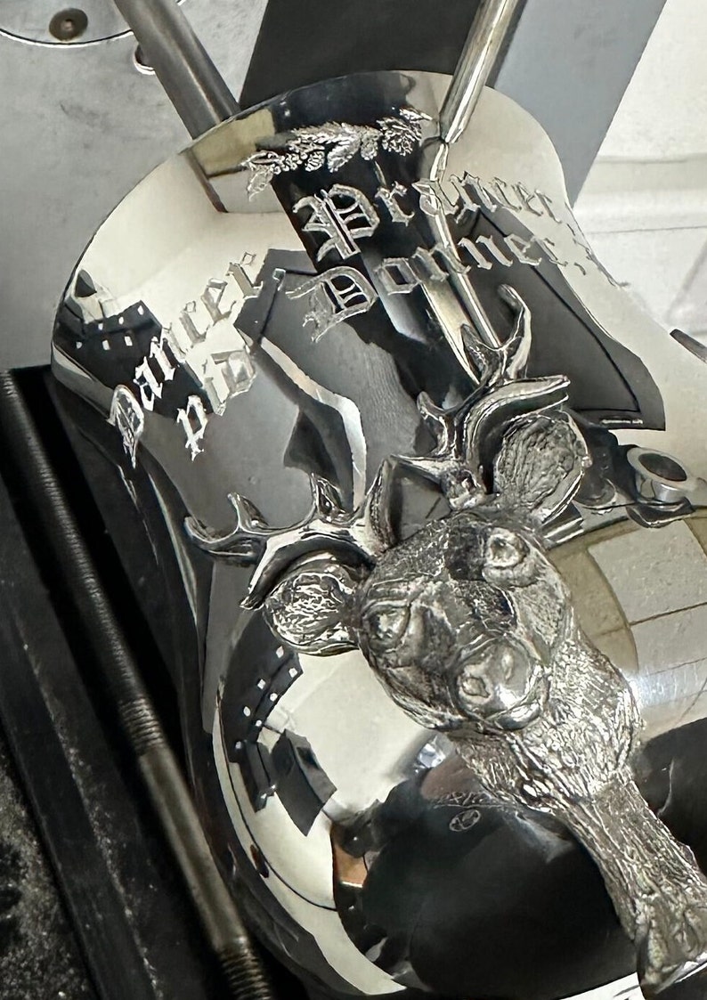 May include: A polished silver-toned metal vessel featuring a three-dimensional deer head relief on the front. The surface is engraved with the text Dancer, Prancer, and Donner in a gothic-style font, accompanied by a small pine cone branch motif.