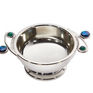 May include: A silver bowl with a circular base and two handles. Each handle features a green and a blue stone. The bowl is made of a shiny metal and has a smooth interior.