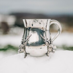 May include: A polished silver-toned metal mug featuring two sculpted deer heads with antlers attached to the base. The mug has a smooth, reflective surface and a curved handle on the right side, showcasing detailed craftsmanship on the animal figures.