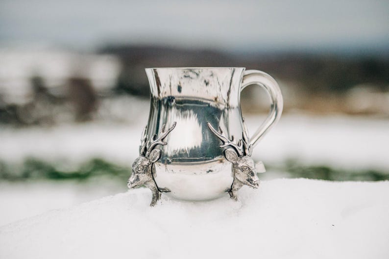 May include: A polished silver-toned metal mug featuring two sculpted deer heads with antlers attached to the base. The mug has a smooth, reflective surface and a curved handle on the right side, showcasing detailed craftsmanship on the animal figures.