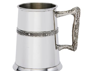 May include: A polished silver-toned metal tankard featuring a smooth cylindrical body with a horizontal band of intricate Celtic knotwork and circular accents. The vessel includes a textured, ornate handle with detailed relief patterns.