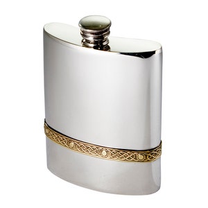 May include: A polished silver-tone metal flask with a curved rectangular body and a screw-top cap. A horizontal band featuring a gold-tone intricate woven knotwork pattern and small circular accents wraps around the lower section of the flask.