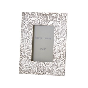 May include: White photo frame with a skull and heart pattern. The frame is designed to hold a 2x3 inch photo. "Photo Frame" and "2"x"3" are printed on the frame.