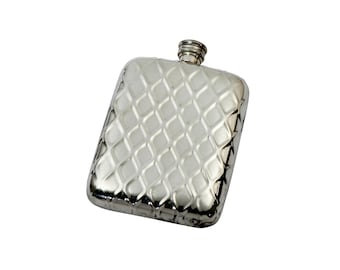 Puede incluir: Una petaca rectangular de metal plateado que presenta un patrón acolchado en forma de diamante en relieve en toda su superficie frontal. La petaca tiene un pequeño tapón de rosca cilíndrico en el centro del borde superior con textura estriada.