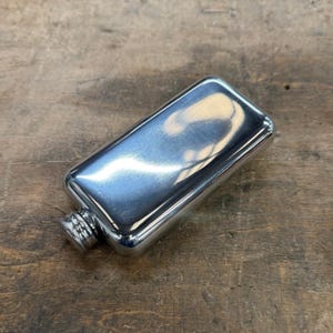 Könnte beinhalten: Eine polierte Edelstahlflasche mit Schraubverschluss. Die rechteckige Flasche hat abgerundete Kanten und eine reflektierende Oberfläche. Die Kappe ist für einen besseren Halt strukturiert. Zum Halten von Flüssigkeiten.