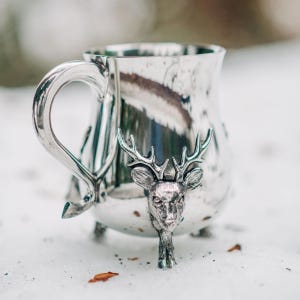 May include: A polished silver-toned metal mug featuring a three-dimensional deer head sculpture on the front. The mug has a curved handle and stands on three small feet, with the deer head extending down to form one of the support legs.