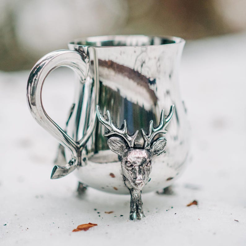 May include: A polished silver-toned metal mug featuring a three-dimensional deer head sculpture on the front. The mug has a curved handle and stands on three small feet, with the deer head extending down to form one of the support legs.