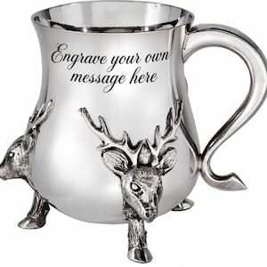 May include: A polished silver-toned metal mug featuring a curved handle and three legs shaped like deer heads with antlers. The front of the mug displays the black text Engrave your own message here, providing a space for personalization.