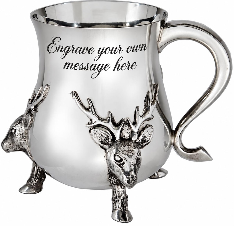 May include: A polished silver-toned metal mug featuring a curved handle and three legs shaped like deer heads with antlers. The front of the mug displays the black text Engrave your own message here, providing a space for personalization.