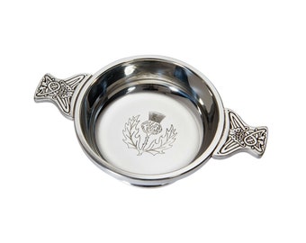 May include: A silver pewter quaich with two handles, featuring a Celtic knot design and a Scottish thistle emblem in the center.