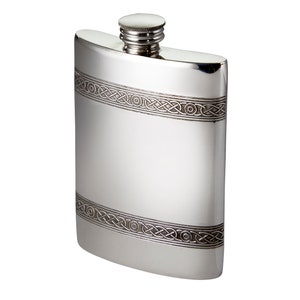 May include: A silver-coloured metal flask with a curved rectangular shape and a screw-top cap. The flask features two horizontal bands of intricate, embossed Celtic knotwork patterns that wrap around the upper and lower sections of the body.